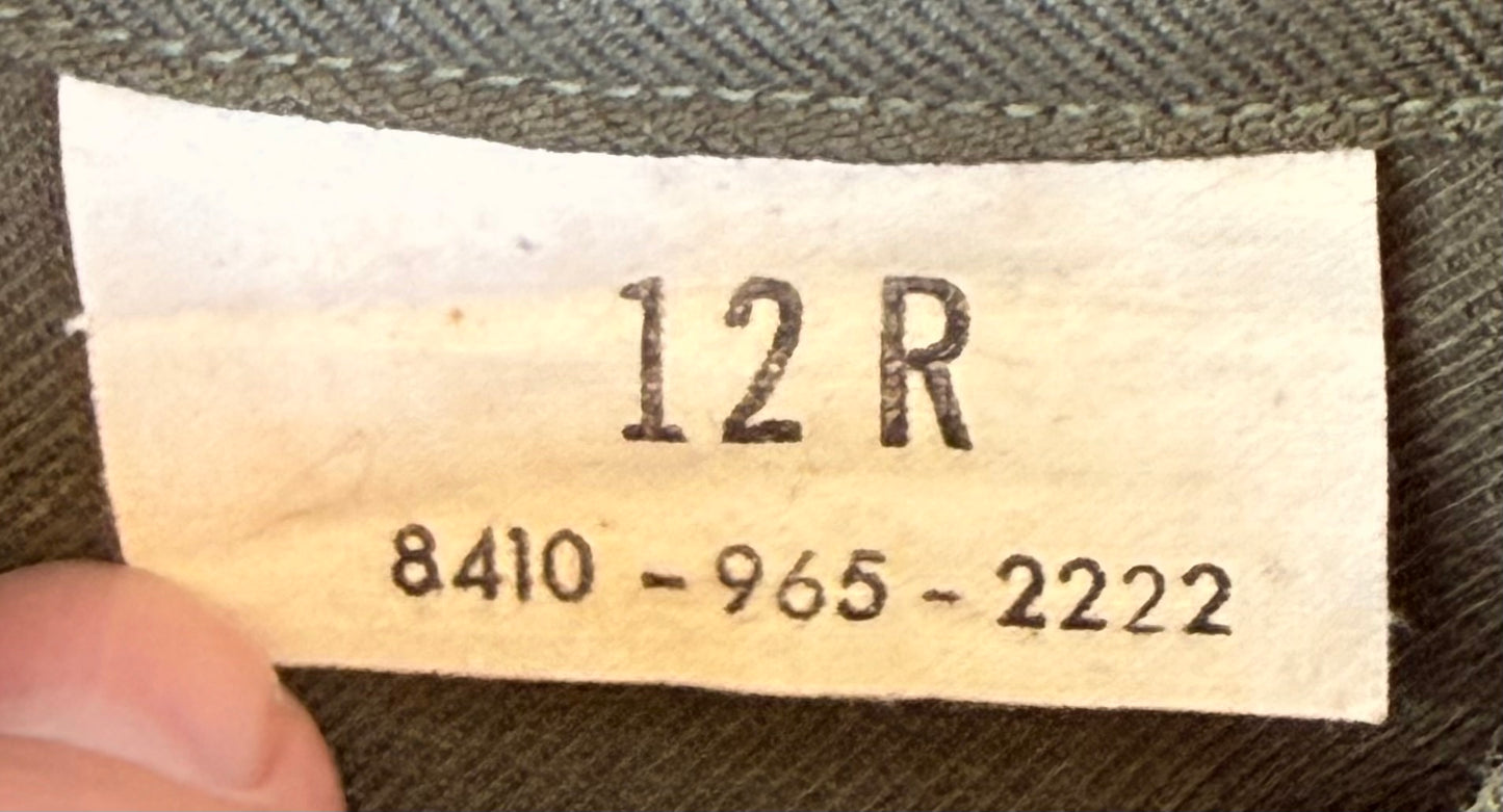 Close-up of a garment size label reading 12R with a serial number, attached to dark olive green fabric and held by a finger.