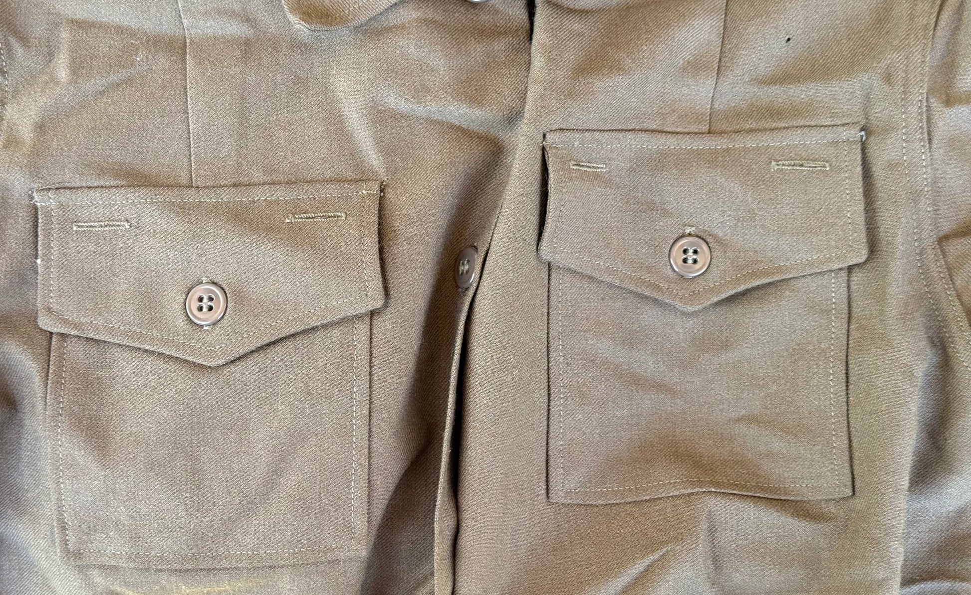 Close-up of a beige or khaki button-up shirt or overshirt showing two front flap pockets with buttons, textured fabric, and part of the button placket.