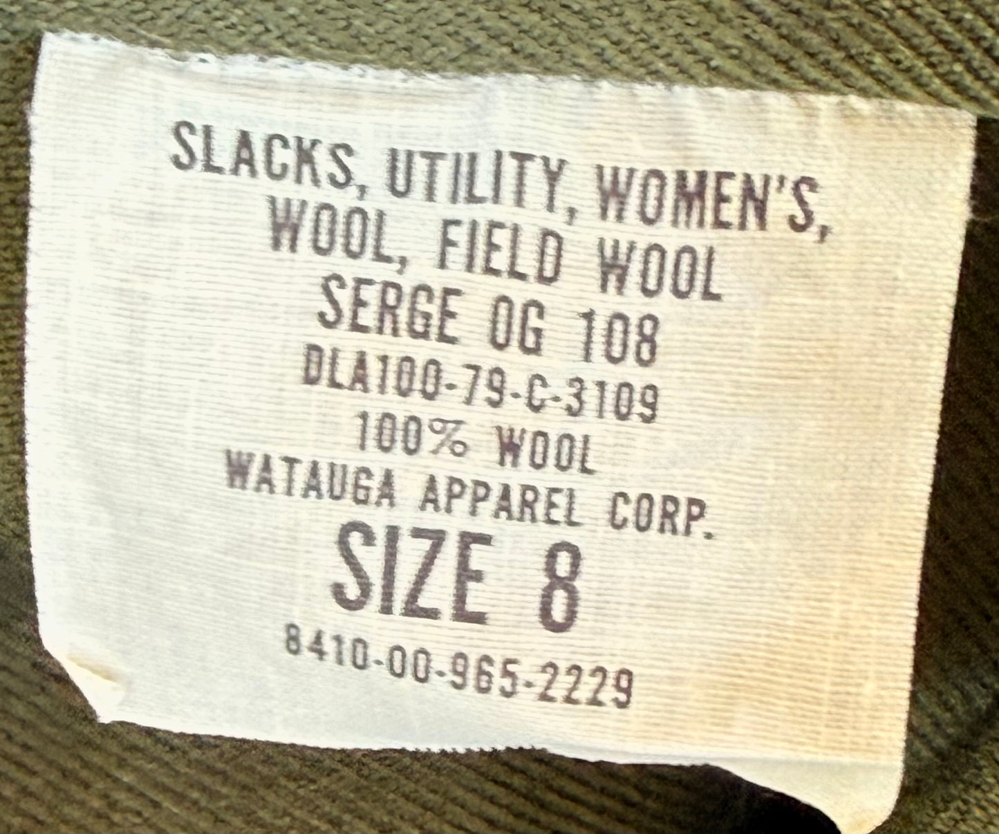 Close-up of a woven fabric label reading 'Slacks, Utility, Women's, Wool, Field Wool SERGE OG 108 DLA100-79-C-3109 100% WOOL WATAUGA APPAREL CORP. SIZE 8' on olive green fabric.