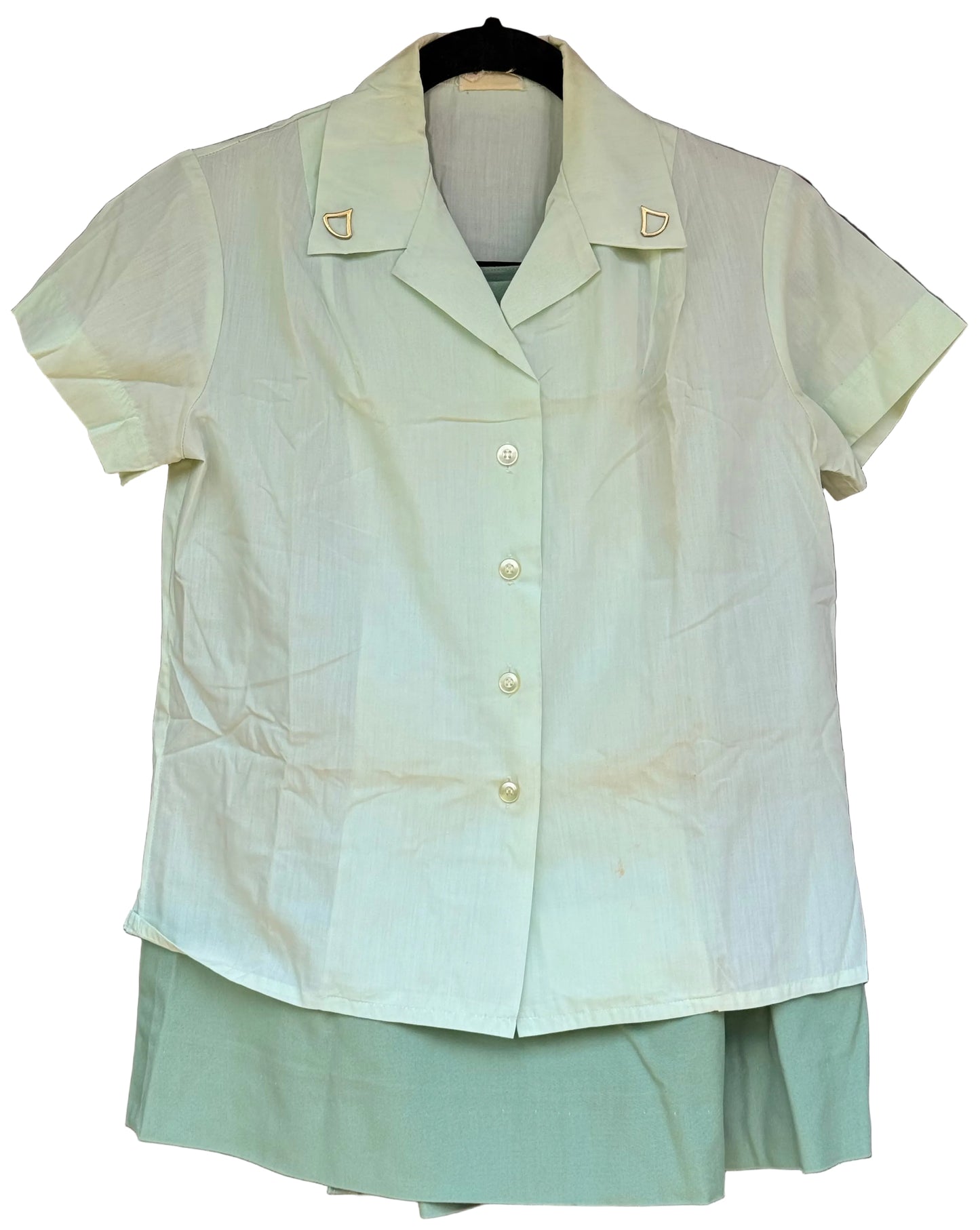 A light sage green short-sleeve button-up shirt with a pointed collar and gold shield-shaped lapel badges, hanging on a black hanger against a dark wood background.