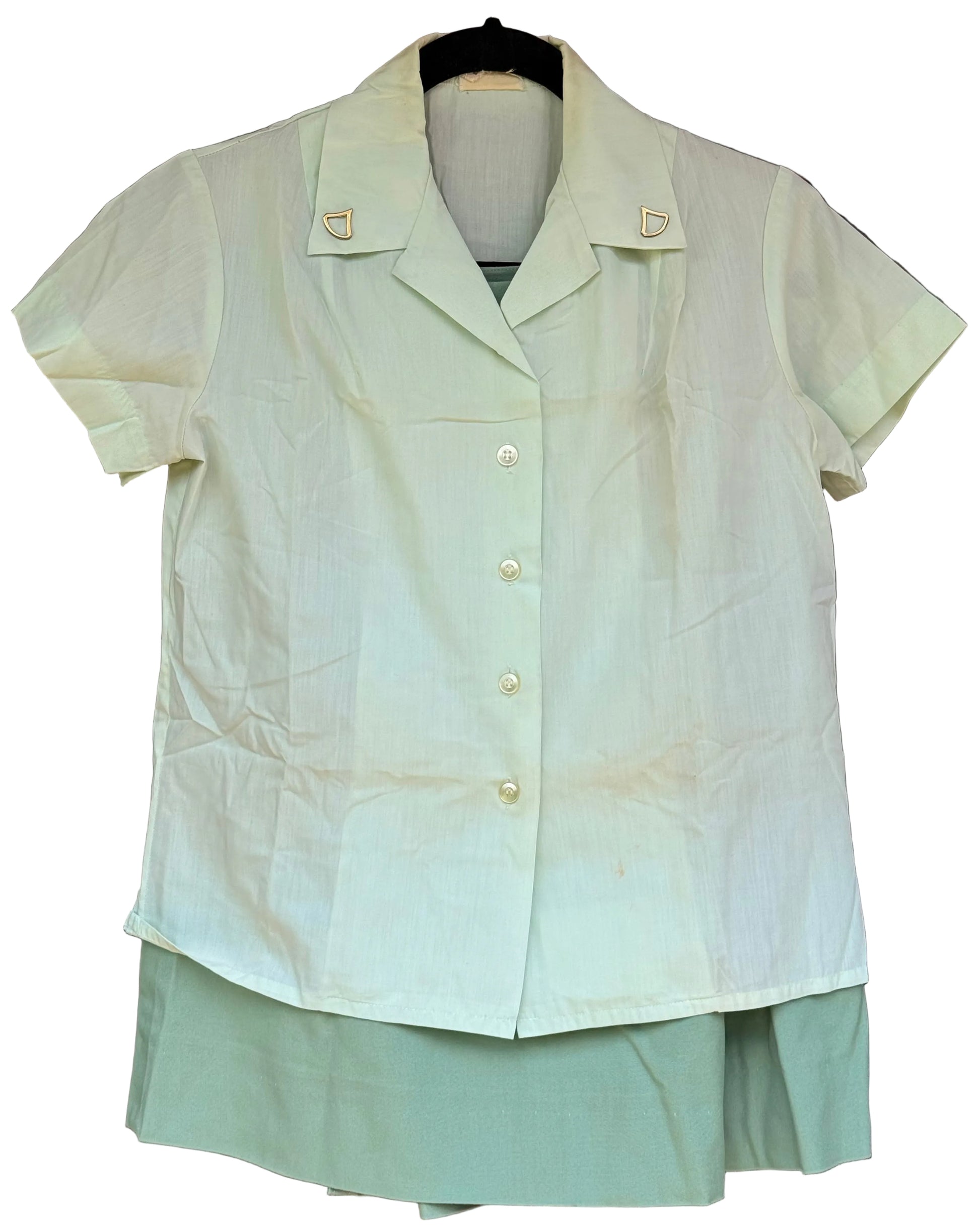 A light sage green short-sleeve button-up shirt with a pointed collar and gold shield-shaped lapel badges, hanging on a black hanger against a dark wood background.