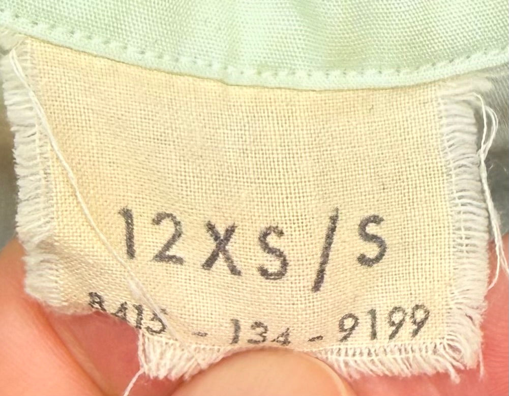 Close-up of a beige fabric size label sewn onto a garment, showing the printed text '12XS/S' and numbers, held by fingers.