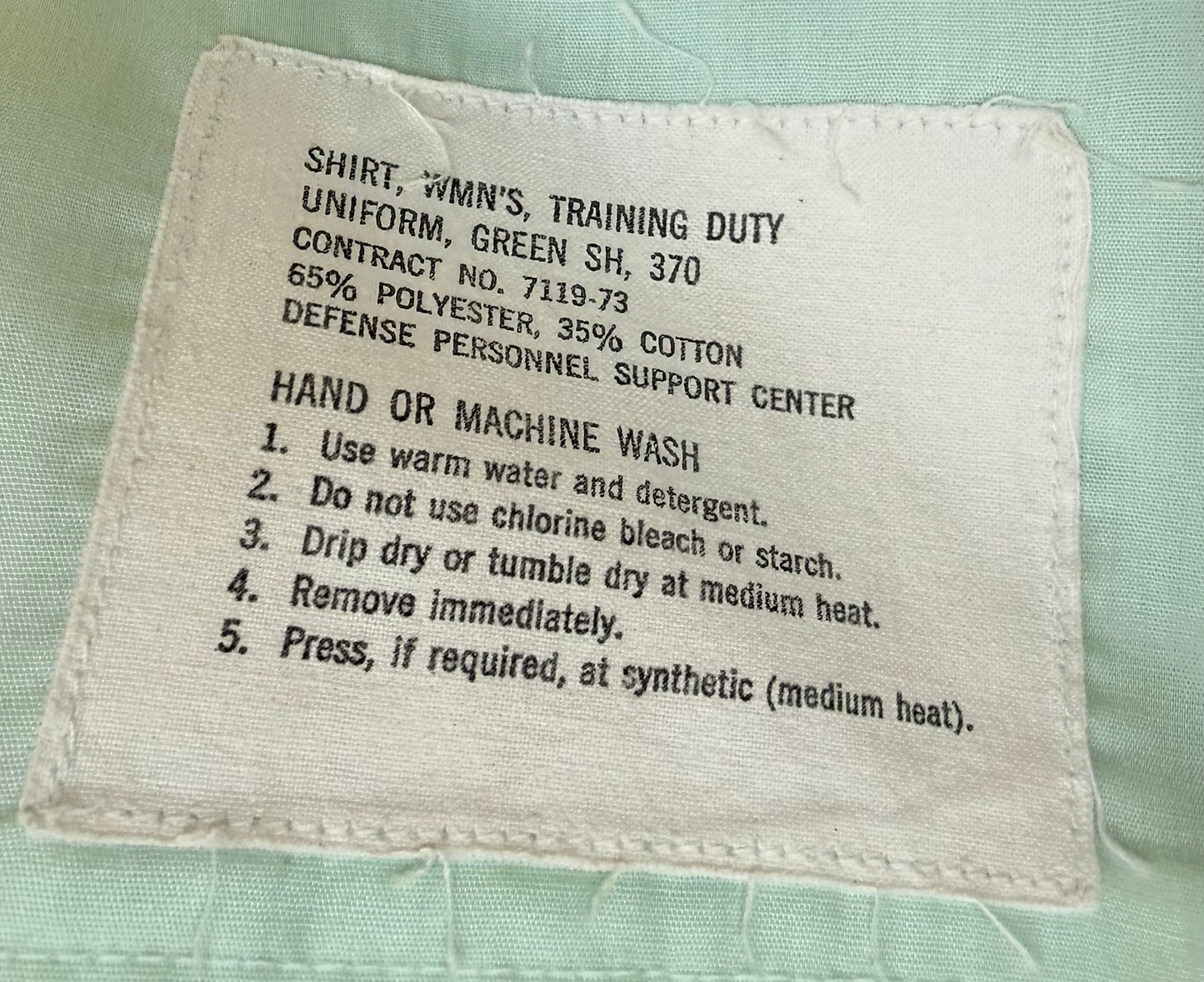 A close-up of a sewn fabric care label on a light green garment. The label reads: 'SHIRT, WMN'S, TRAINING DUTY UNIFORM, GREEN SH, 370 CONTRACT NO.
