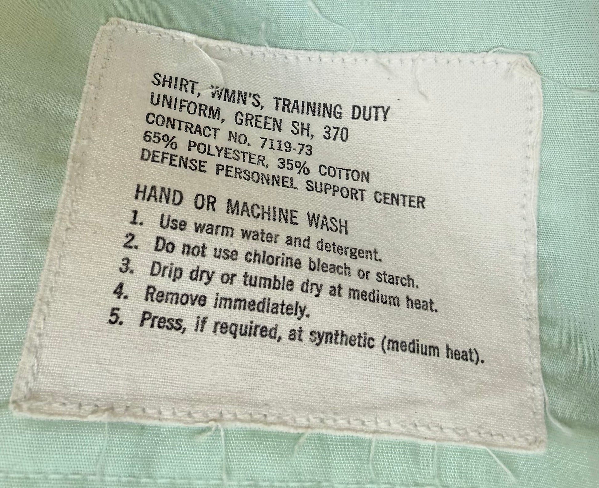 A close-up of a sewn fabric care label on a light green garment. The label reads: 'SHIRT, WMN'S, TRAINING DUTY UNIFORM, GREEN SH, 370 CONTRACT NO.