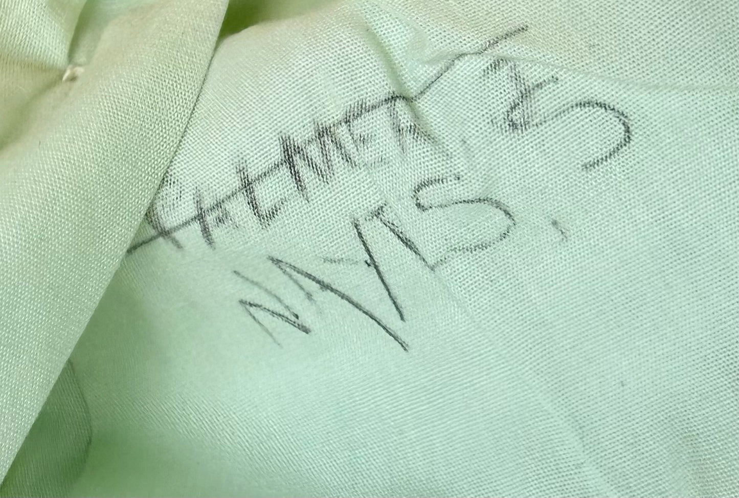 A close-up of pale mint green fabric with handwritten black ink markings reading 'PATMAN' and 'NYS 2'.