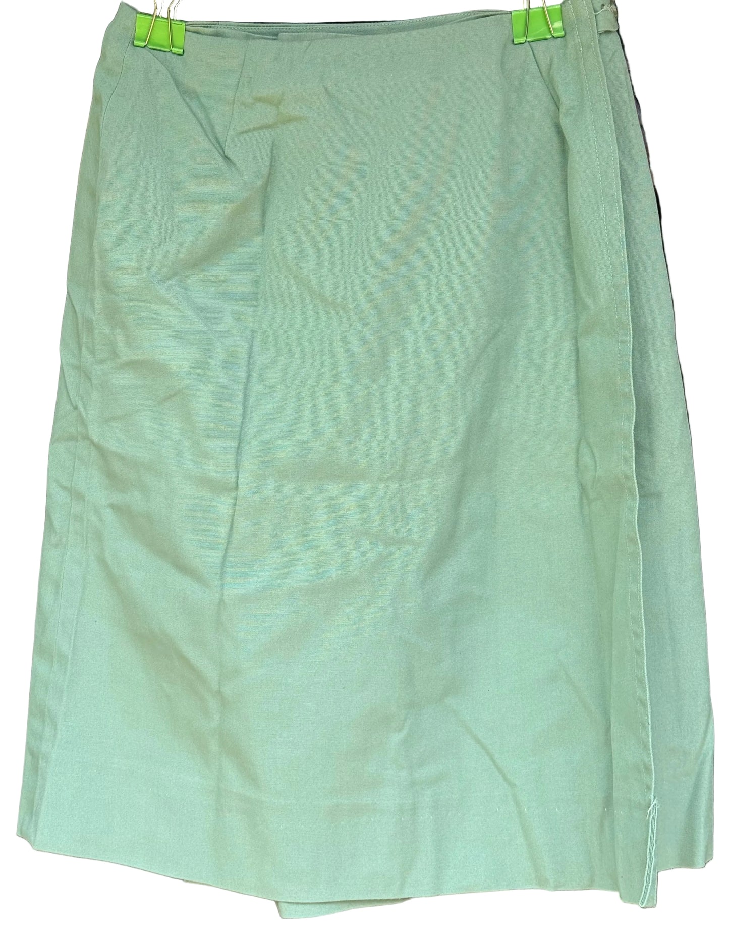 A light sage green skirt with a subtle textured pattern, hanging on a black hanger with bright green clips against a dark wooden background.