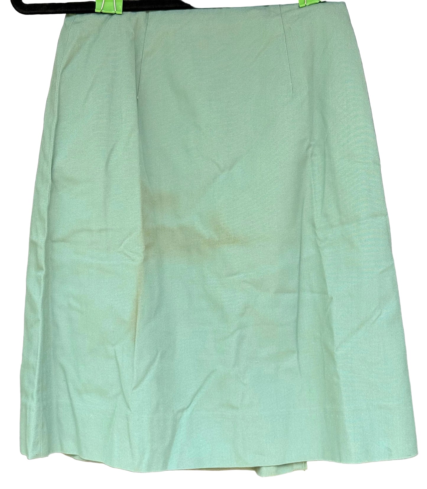 A light sage green skirt with a subtle textured pattern, hanging on a black hanger with green clips against a dark brown carved wooden background.