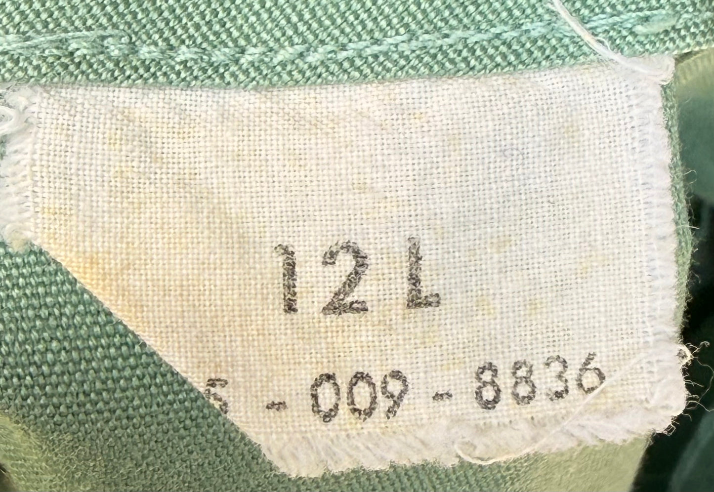 Close-up of a fabric label sewn onto a green woven textile, showing the size marking '12 L' and a serial number.