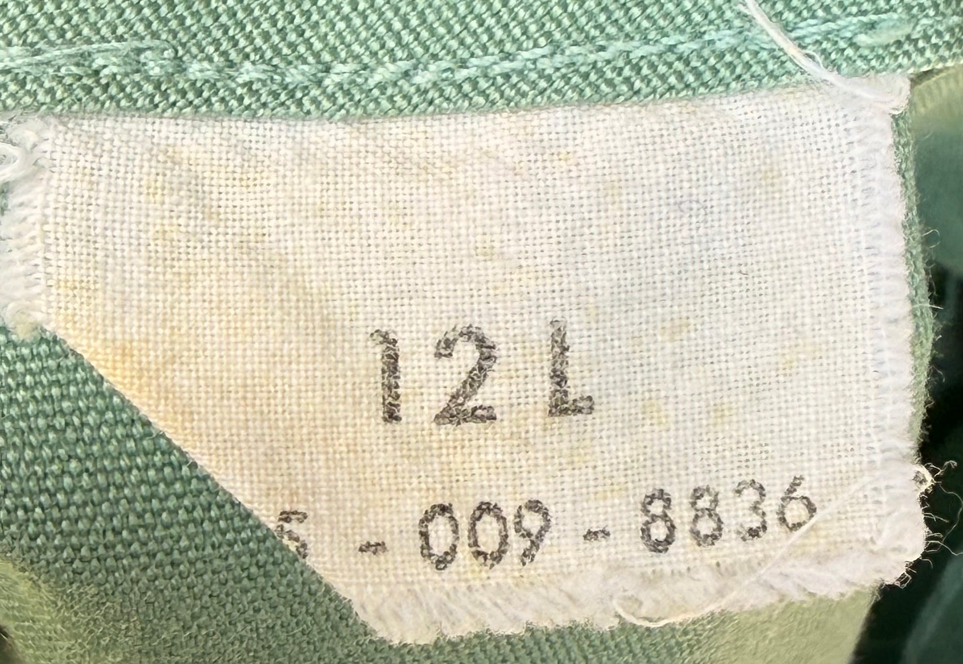 Close-up of a fabric label sewn onto a green woven textile, showing the size marking '12 L' and a serial number.