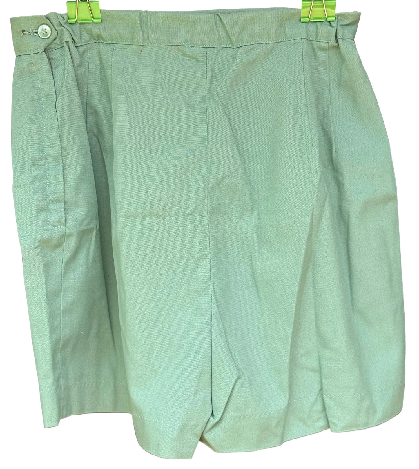 A light sage green pleated skirt or shorts with a button closure at the waist, hanging on a black hanger with green clips against a dark wood background.