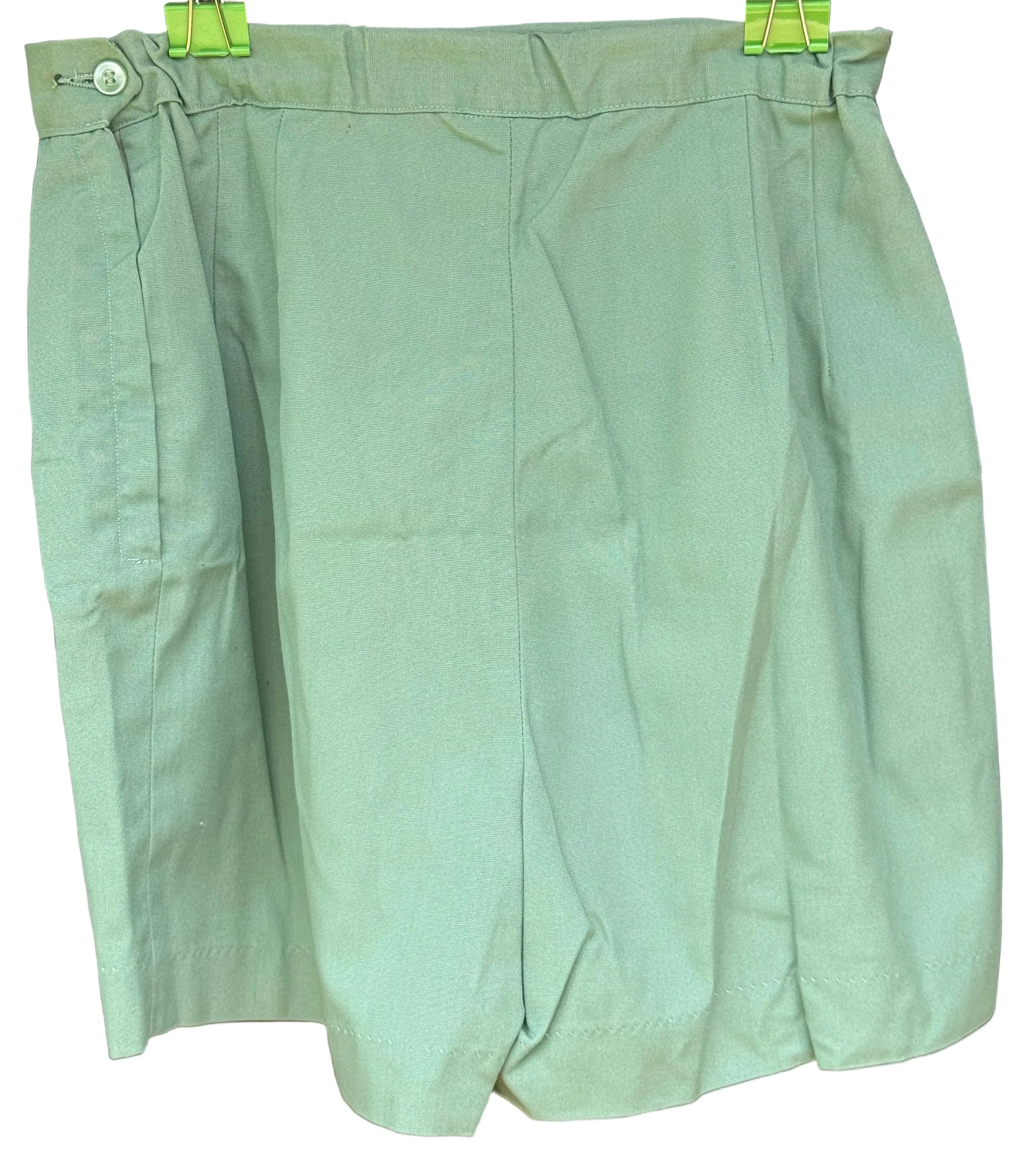 A light sage green pleated skirt or shorts with a button closure at the waist, hanging on a black hanger with green clips against a dark wood background.