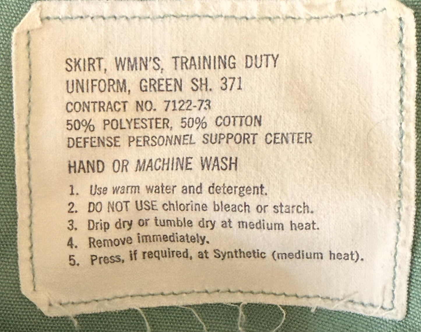 A close-up of a vintage uniform label sewn onto green fabric. The label reads 'SKIRT, WMN'S, TRAINING DUTY UNIFORM, GREEN SH.
