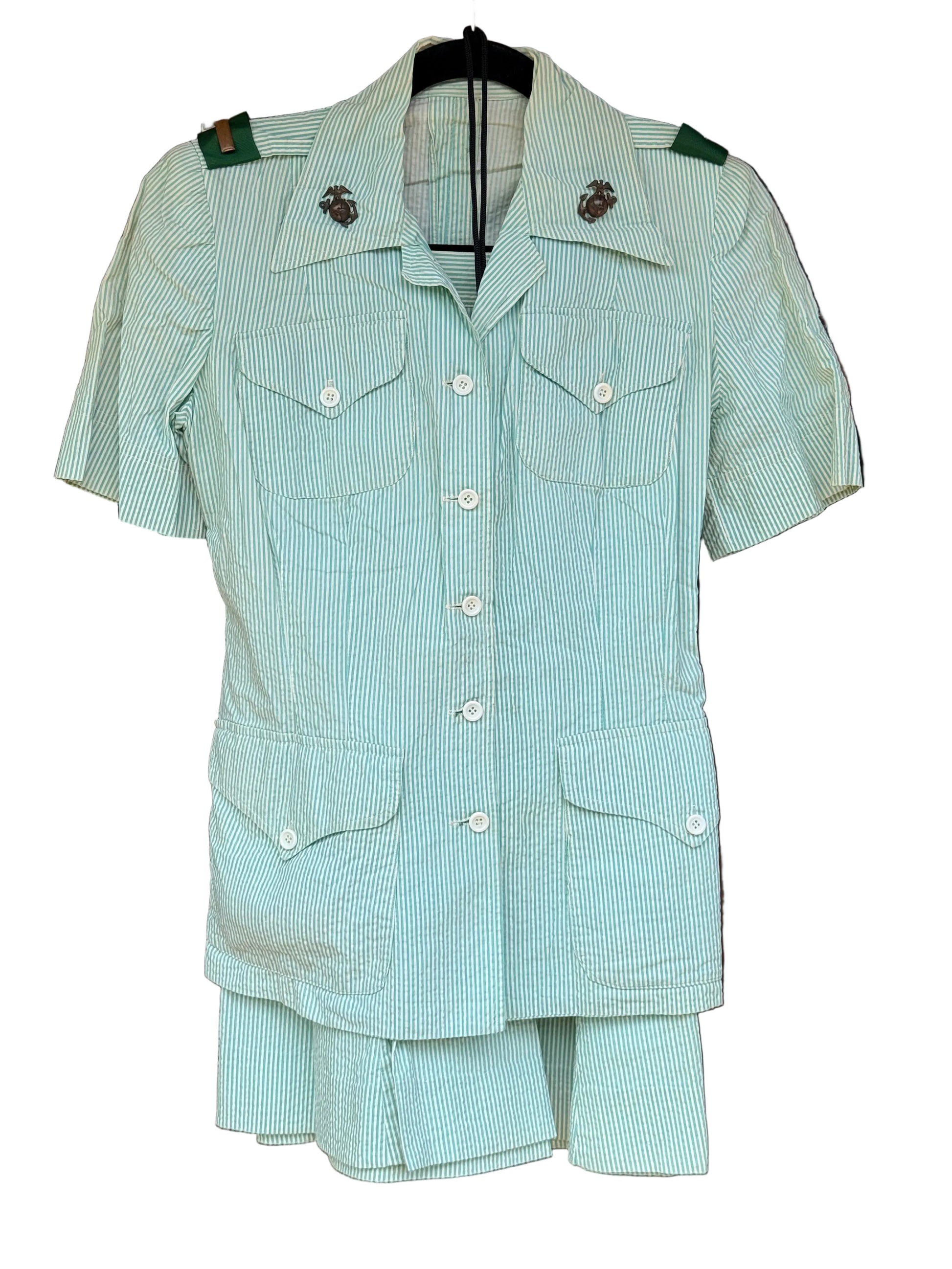 A short-sleeve military-style shirt jacket and matching shorts set in a pale mint green and white pinstripe fabric, hanging on a black hanger against a dark wood background.