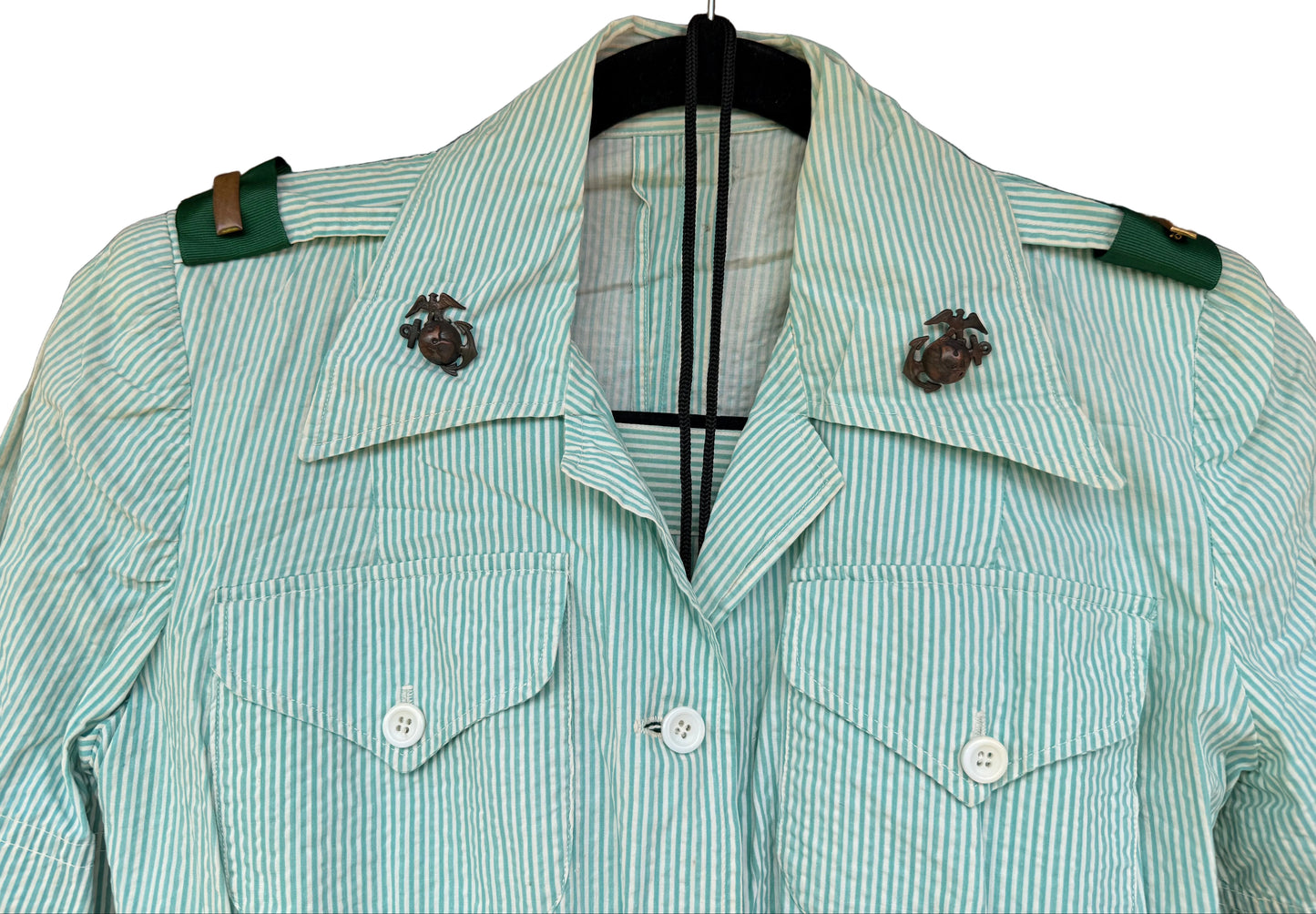 A light green and white vertically striped military-style jacket with green shoulder tabs and brass-colored Marine Corps EGA emblems on the collar, hanging on a black hanger against a dark wood background.