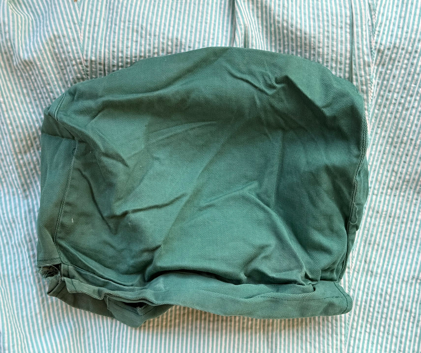 A dark green fabric cover or pouch with a gathered, elasticized bottom, laid flat on a light blue and white striped fabric background.