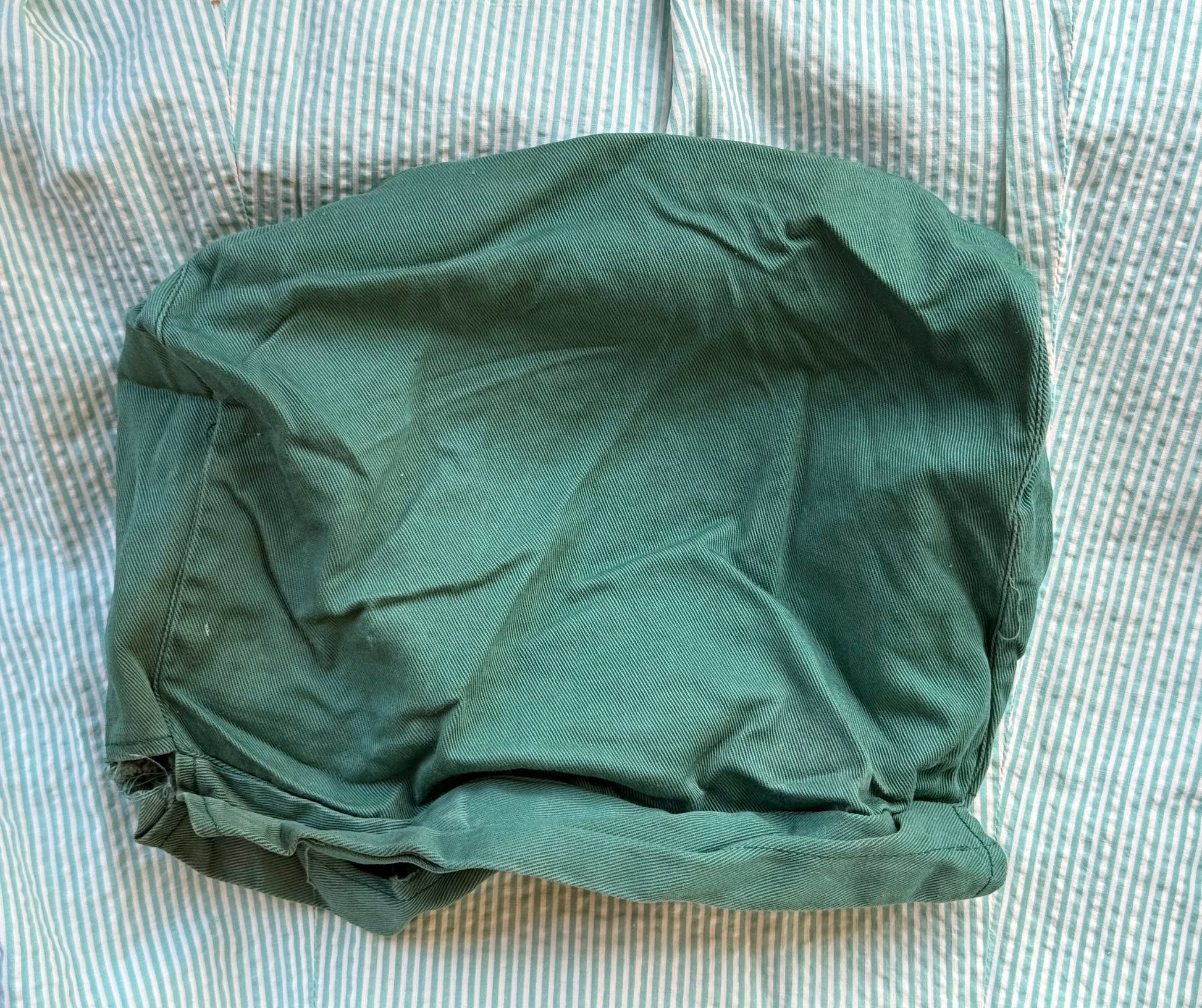 A dark green fabric cover or pouch with a gathered, elasticized bottom, laid flat on a light blue and white striped fabric background.