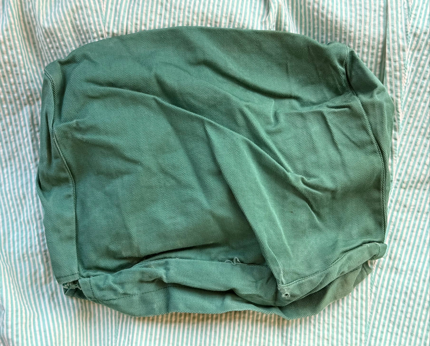 An olive green fabric pouch or cover with a matte finish, laid flat on a white and blue striped fabric background.