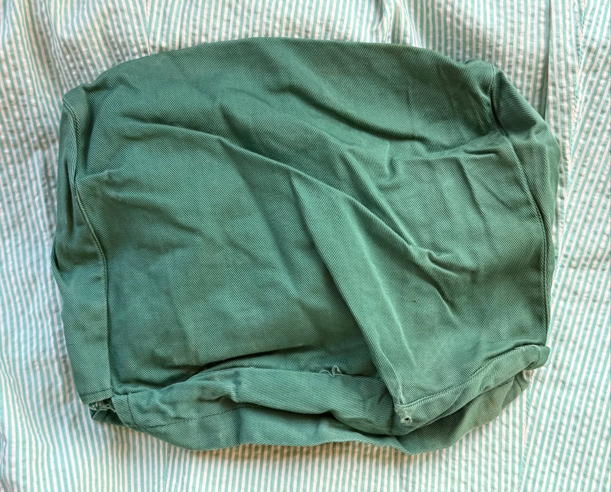 An olive green fabric pouch or cover with a matte finish, laid flat on a white and blue striped fabric background.