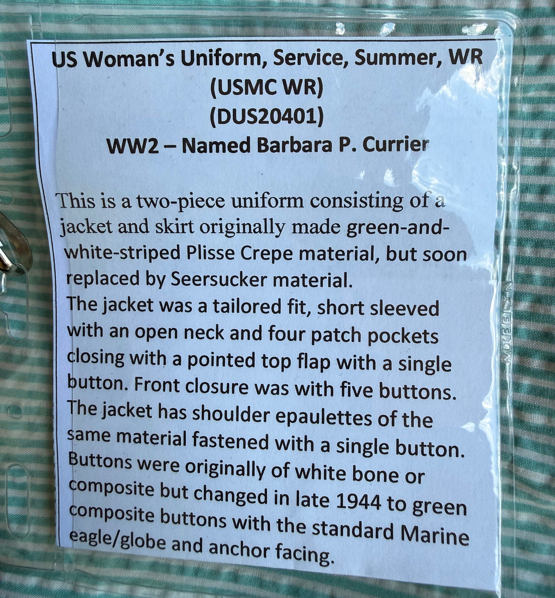 A printed paper label showing historical product details for US Woman's Uniform WW2 Size Barbarella P. Carrier, photographed on a green and white striped fabric surface.