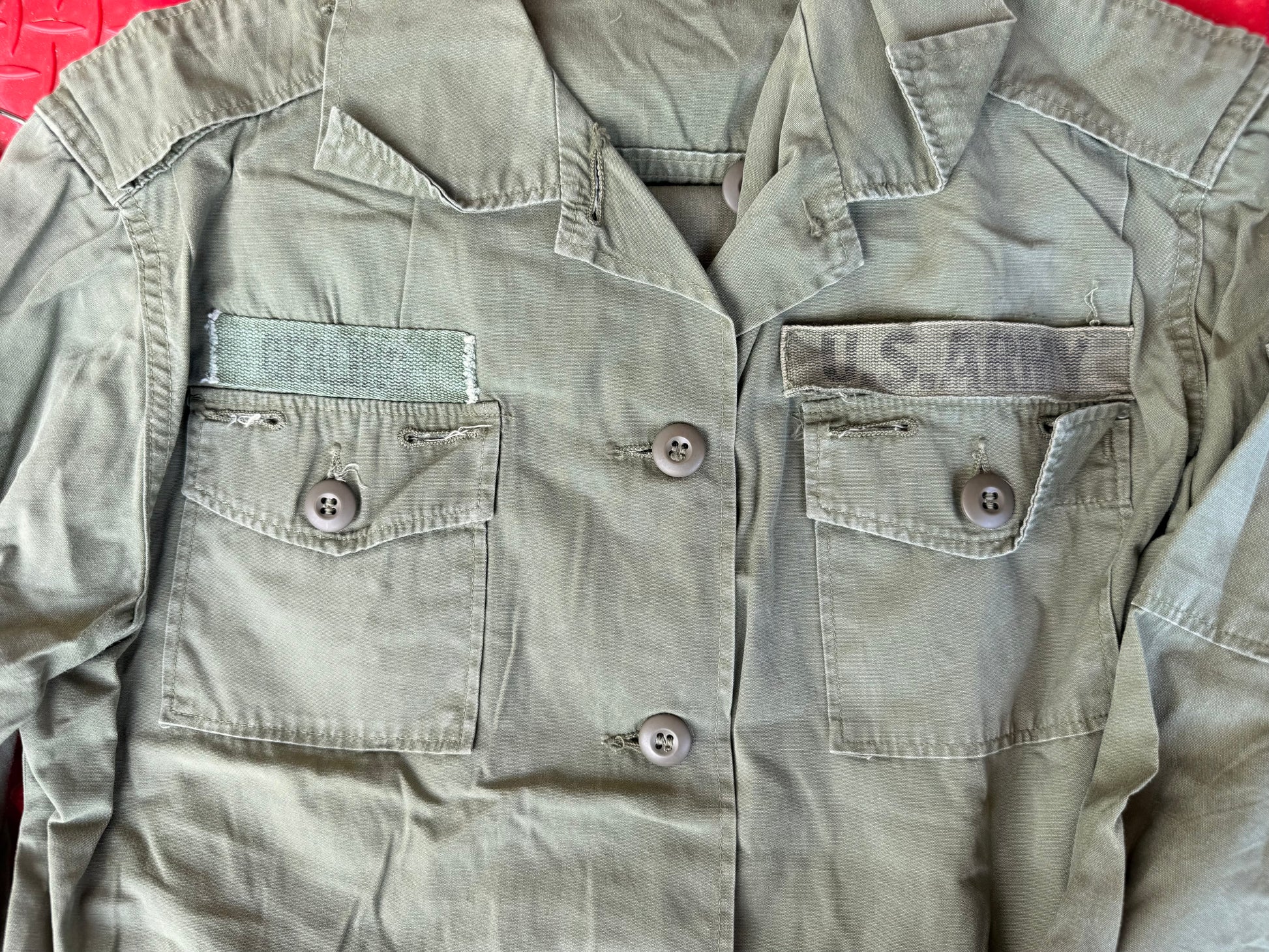 A close-up of a military-style utility jacket in olive drab green, featuring two buttoned chest pockets with 'U.S.