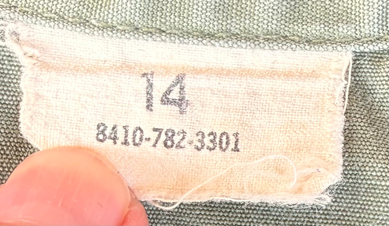 Close-up of a fabric size label reading 14 and 8410-782-3301, held by a finger against olive green woven fabric.
