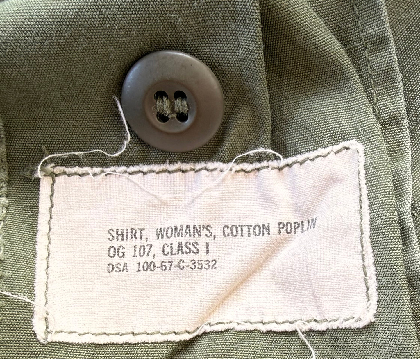 Close-up of an olive green cotton poplin shirt label sewn onto the inside collar, with a dark gray button above it. The label reads 'SHIRT, WOMAN'S, COTTON POPLIN OG 107, CLASS I DSA 100-67-C-3532'.
