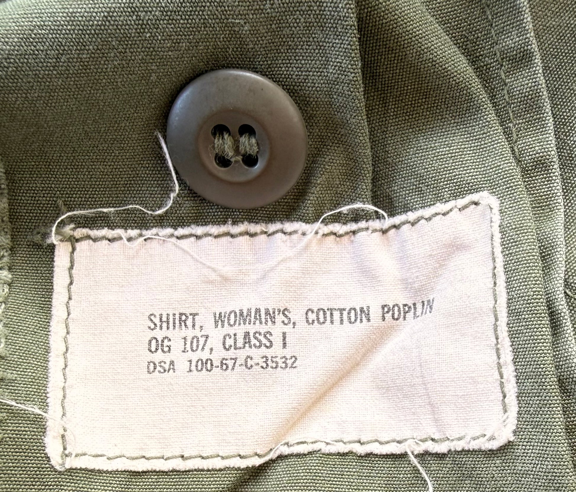 Close-up of an olive green cotton poplin shirt label sewn onto the inside collar, with a dark gray button above it. The label reads 'SHIRT, WOMAN'S, COTTON POPLIN OG 107, CLASS I DSA 100-67-C-3532'.