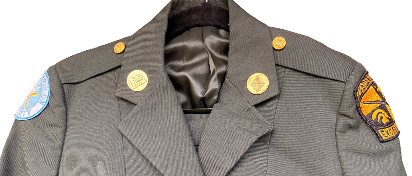 A military-style olive drab uniform jacket with gold buttons and shoulder insignia, displayed on a hanger against a wooden background.