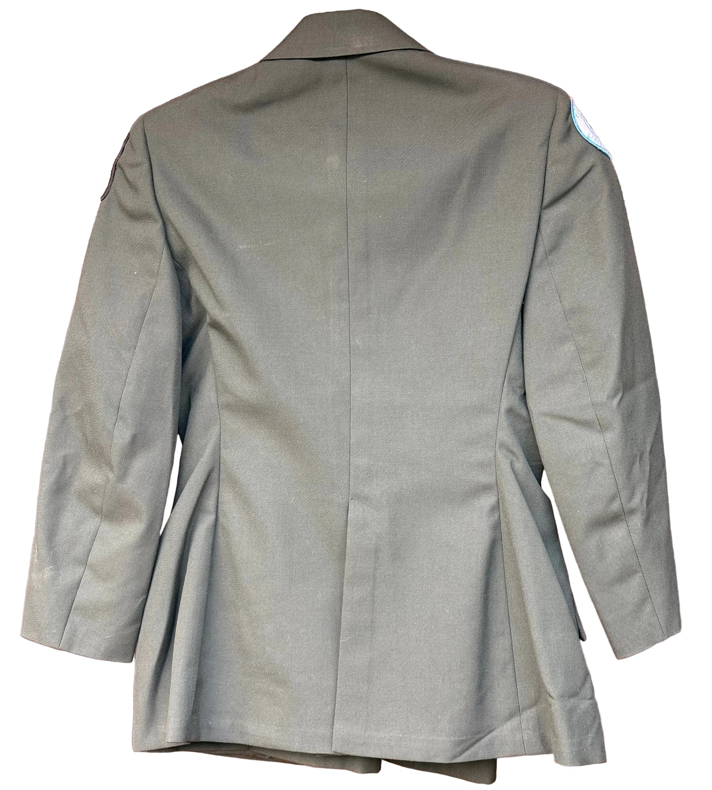 An olive green military-style blazer shown from the back, hanging against a carved reddish-brown wooden background.