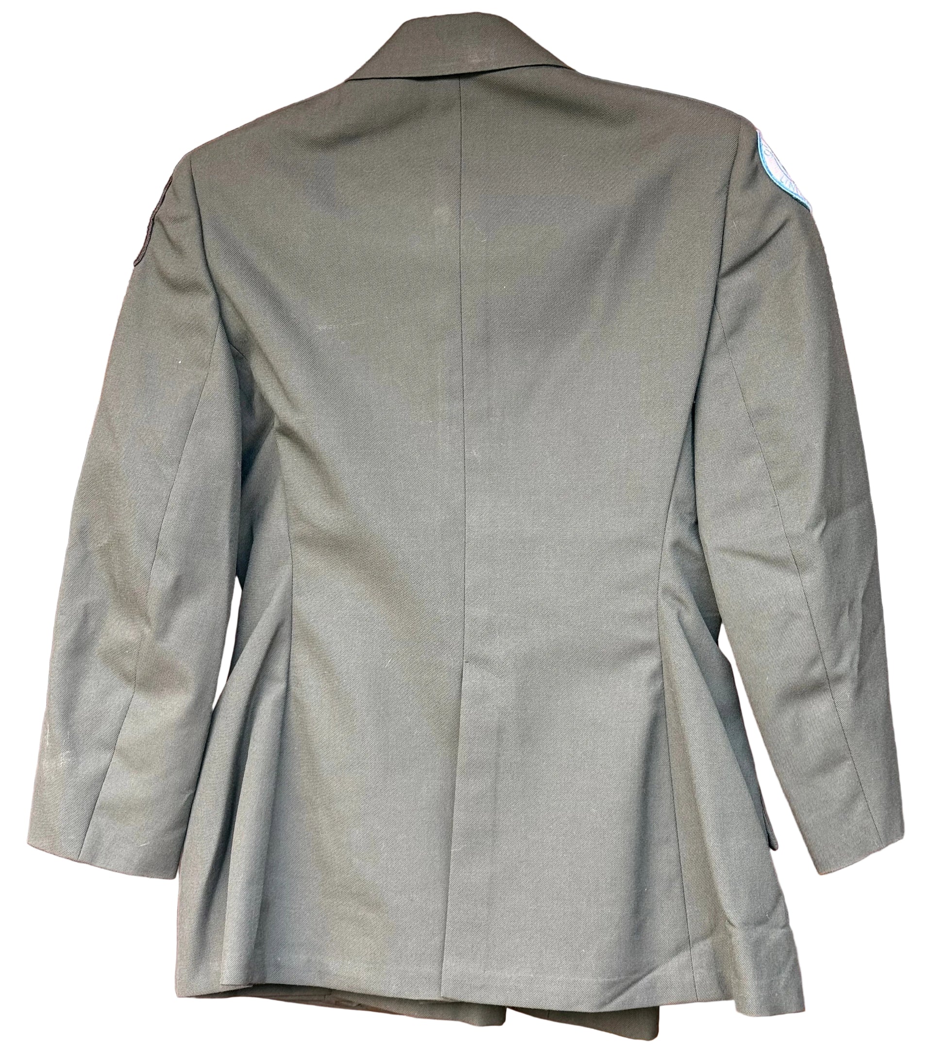 An olive green military-style blazer shown from the back, hanging against a carved reddish-brown wooden background.