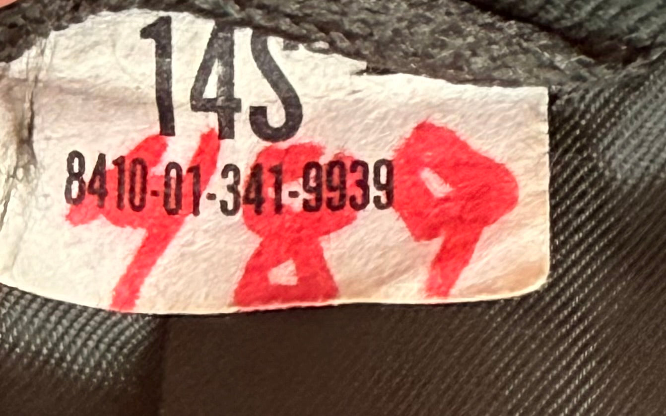 Close-up of a garment interior label showing the size code 14S, a printed serial number, and red handwritten price markings on a dark textured fabric background.