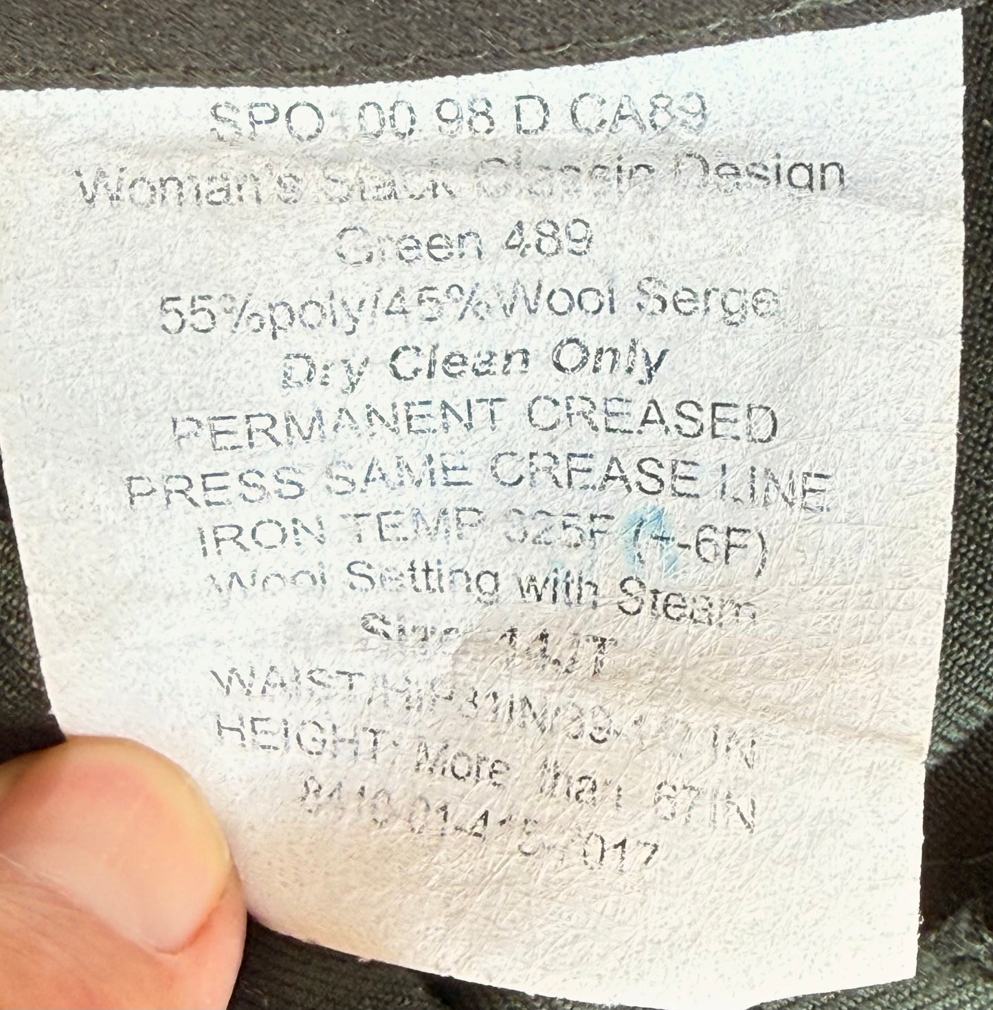 A close-up of a garment's sewn-in care label held by a finger, showing text for 'Woman's Stack Classic Design Green 489' with wool and polyester composition and care instructions.