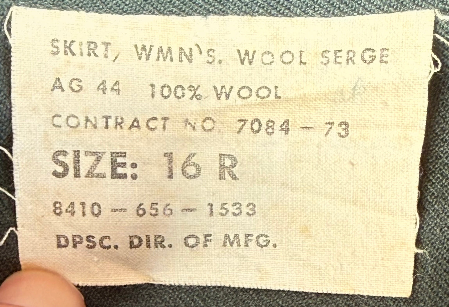 A close-up of a vintage beige fabric label sewn onto a dark gray garment. The label reads 'SKIRT, WMN'S WOOL SERGE AG 44 100% WOOL CONTRACT NO.