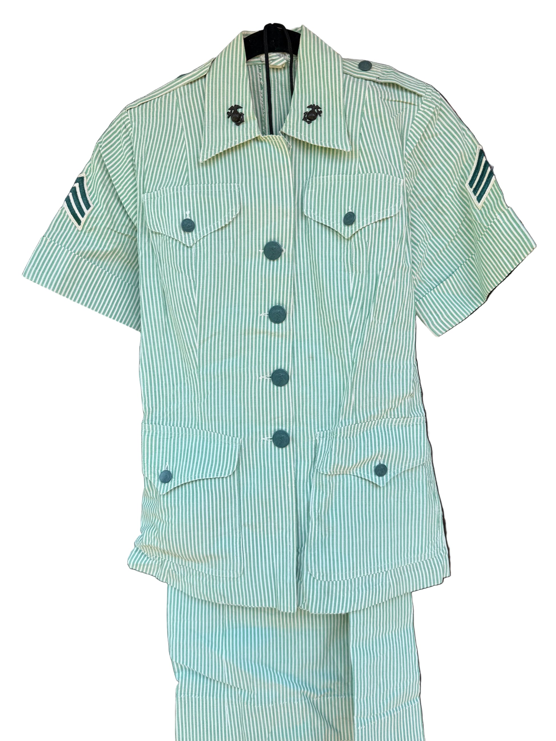 A short-sleeve military-style shirt and matching trousers set in a pale green and white vertical stripe pattern, hanging on a black hanger against a dark wooden background.