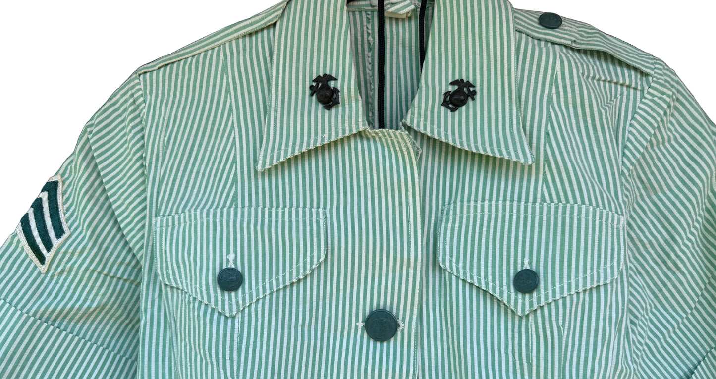 A short-sleeve military-style uniform shirt with vertical green and white stripes, featuring a pointed collar with black insignia, button-front closure, and chest flap pockets, shown hanging against a dark wood background.