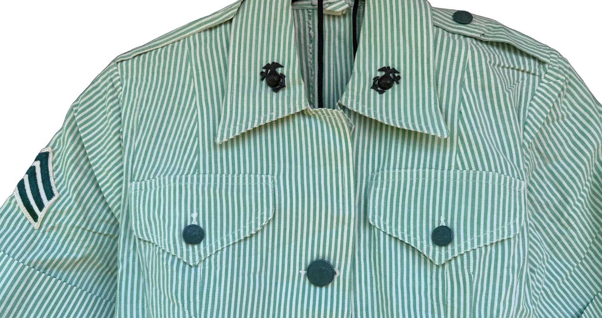 A short-sleeve military-style uniform shirt with vertical green and white stripes, featuring a pointed collar with black insignia, button-front closure, and chest flap pockets, shown hanging against a dark wood background.