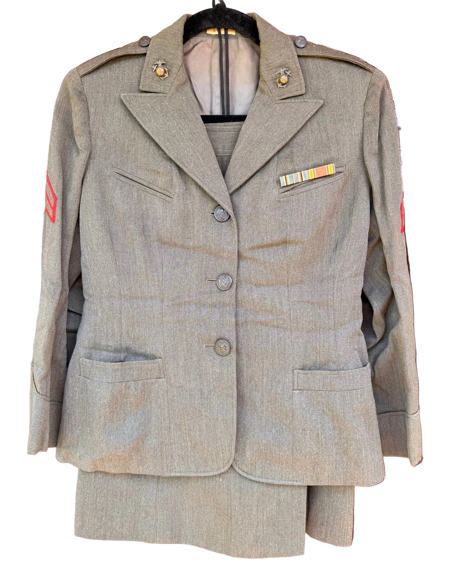 A military-style wool uniform jacket and matching skirt set in a heathered khaki-beige fabric, hanging on a black hanger against a carved dark wood background. The jacket features shoulder epaulets, a three-button front, flap pockets, and a small service ribbon on the chest.
