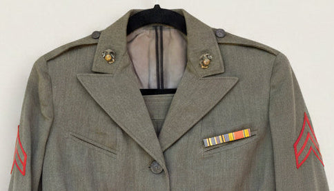 A close-up of a military-style olive drab jacket displayed on a mannequin or hanger, featuring shoulder insignia, chest pockets, a striped service ribbon on the left chest, and a red sleeve tab.