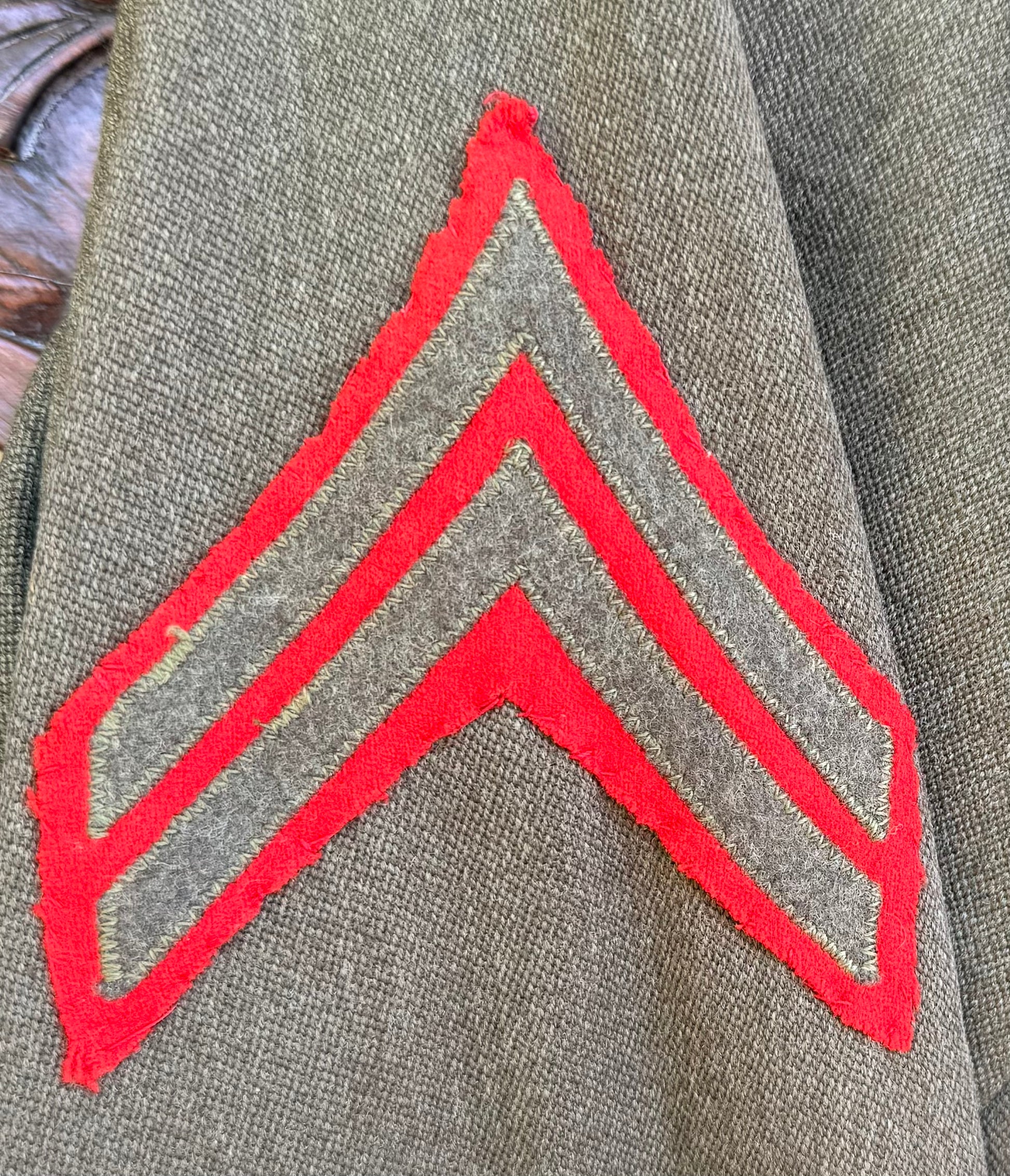 A close-up of a military-style chevron rank insignia on an olive drab jacket. The insignia features two bright red stripes on a gray background, set against the textured olive drab fabric of the uniform.