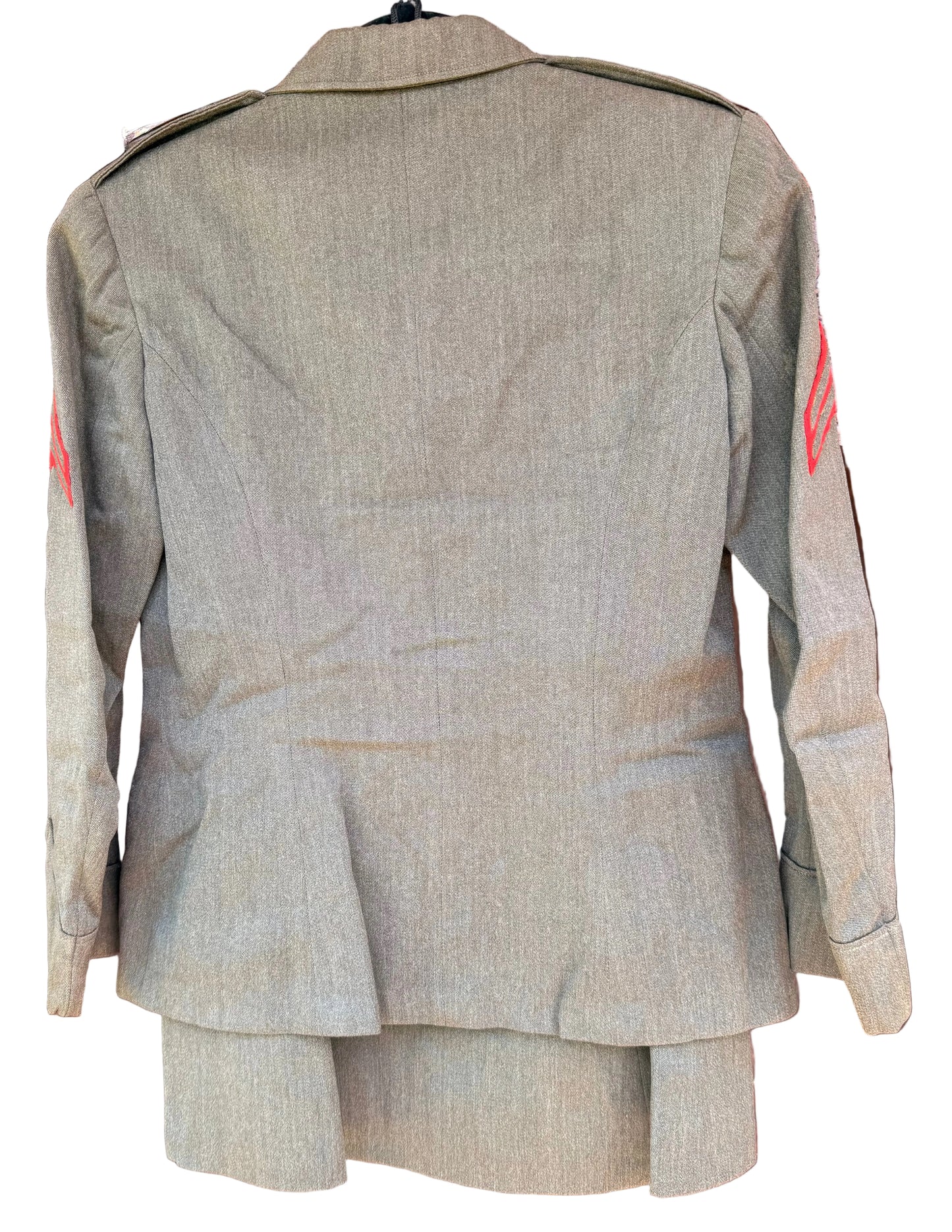 A military-style olive drab uniform jacket shown from the back, hanging against a carved wooden background. The jacket features shoulder epaulets and red insignia on the upper sleeves.