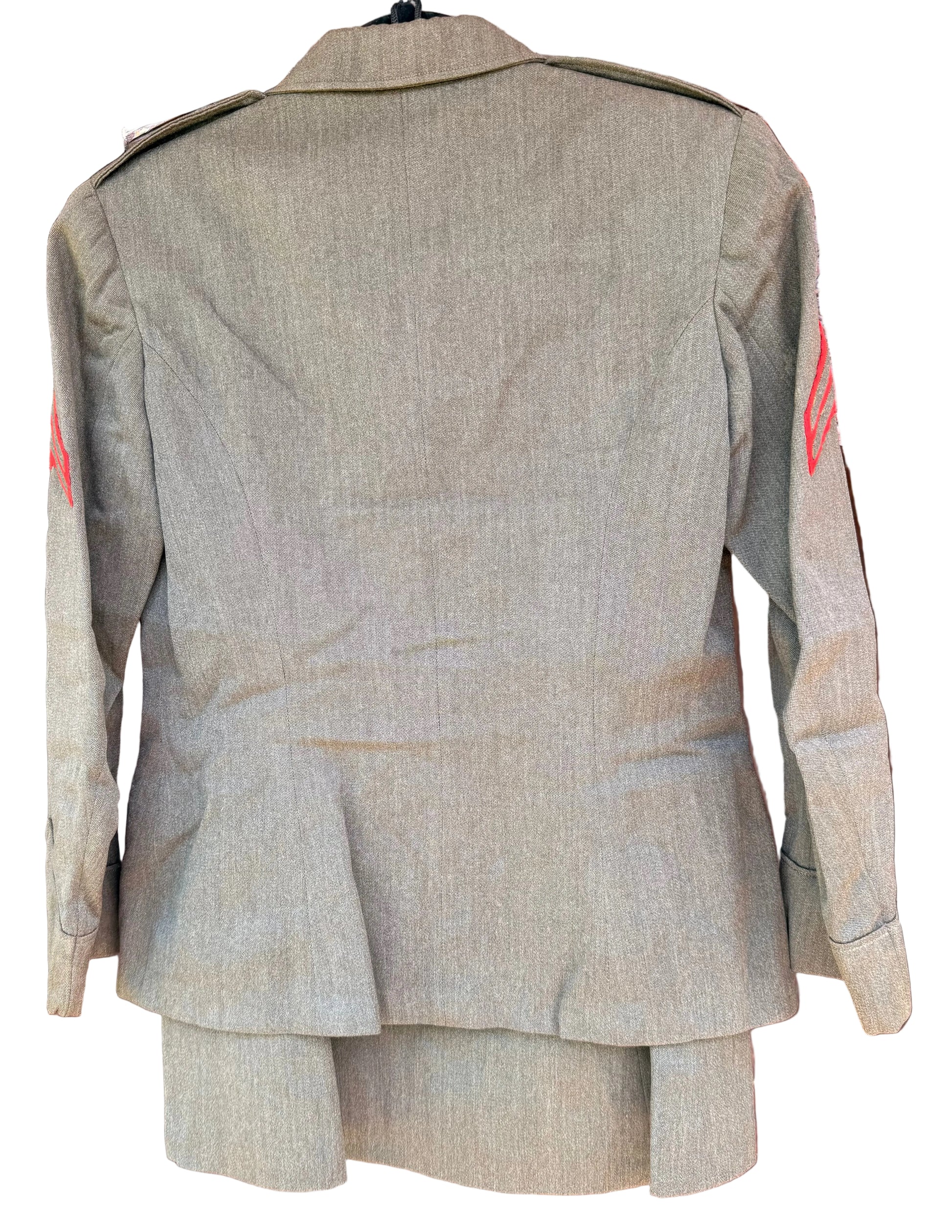 A military-style olive drab uniform jacket shown from the back, hanging against a carved wooden background. The jacket features shoulder epaulets and red insignia on the upper sleeves.