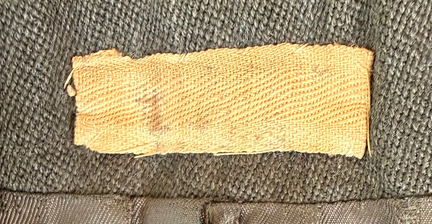 A close-up of a rectangular, tan woven fabric patch or label sewn onto a dark gray textured fabric background.