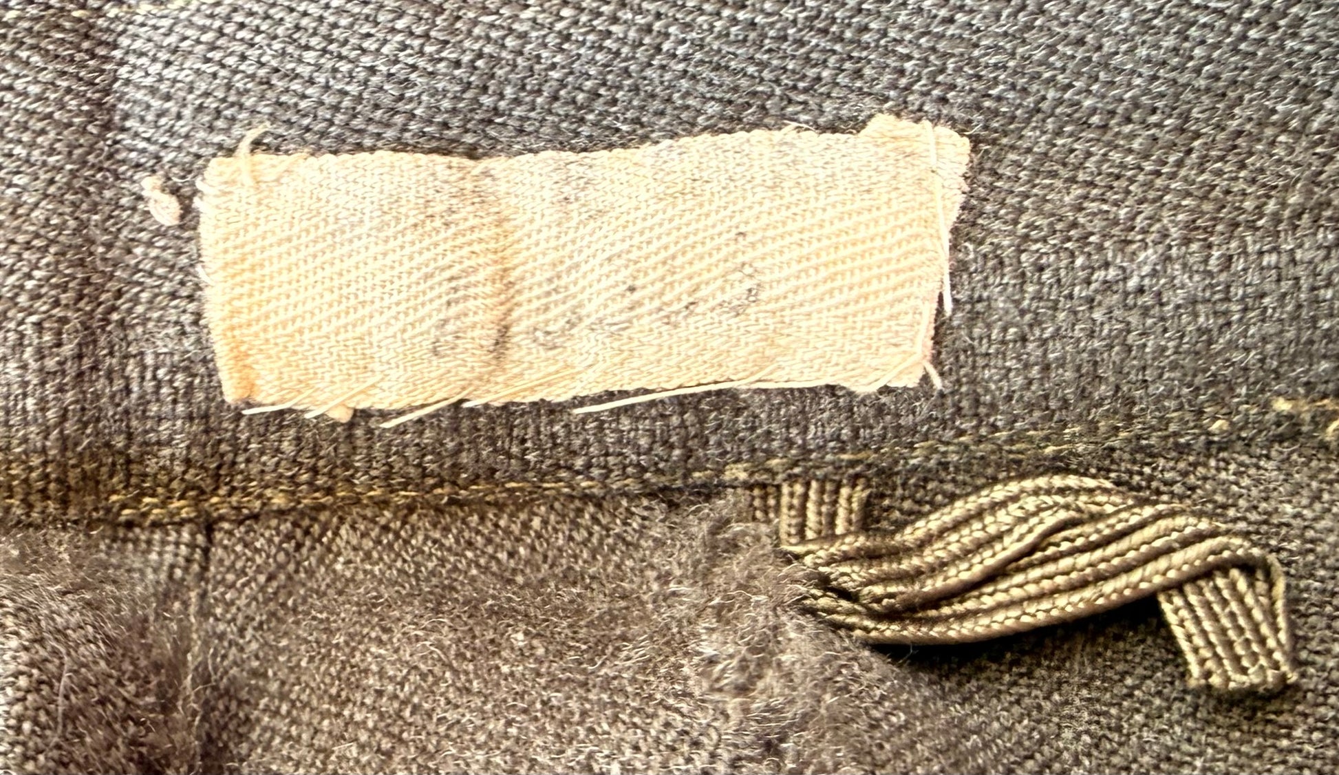 Close-up of a vintage brown woven textile, likely a jacket or coat, showing a light beige fabric label sewn onto the inside and a matching brown woven tie closure.